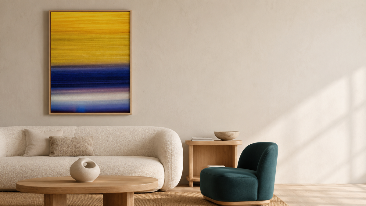 A framed abstract wall art print hangs above a cream-colored sofa in a modern living room. The artwork features horizontal bands of yellow, blue, and white, and is displayed in a light wood frame.